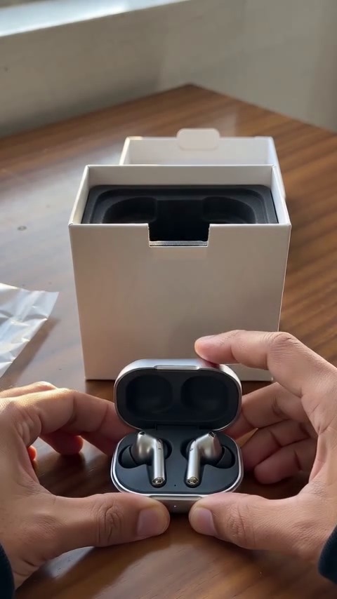 Medium-brown hands open a white earbuds box and reveal a silver charging case on a walnut table